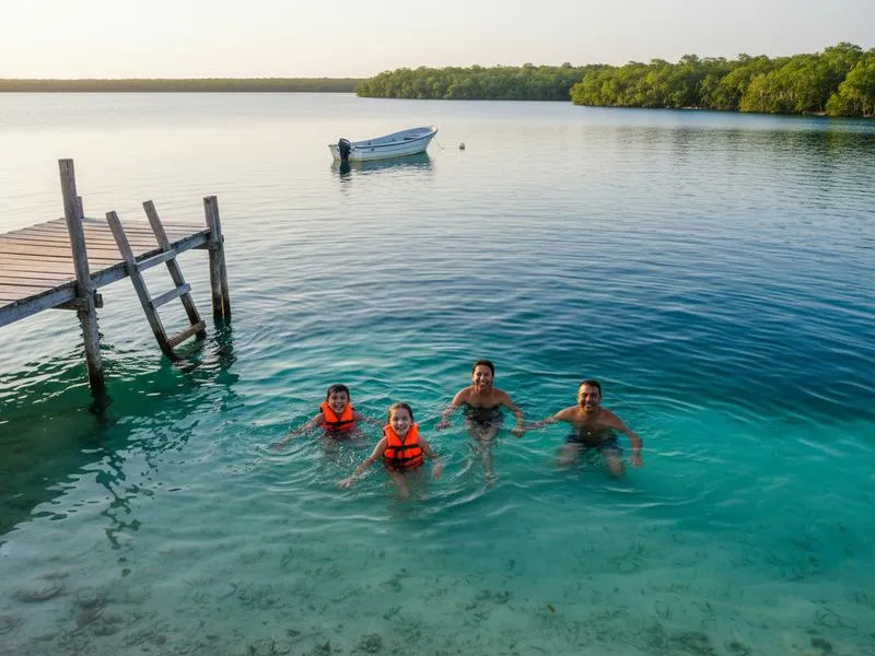 Family swimming in the brilliant blue-green freshwater Bacalar Lagoon in Quintana Roo Mexico, known as the Lake of Seven Colors, with wooden dock and palm trees in background