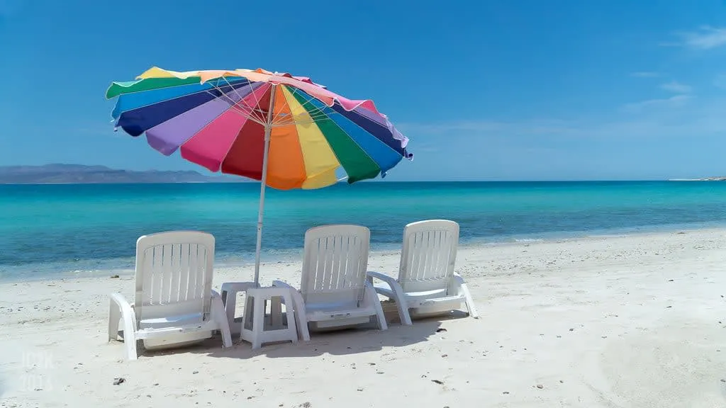 Three white beach chairs under a rainbow umbrella on white sand