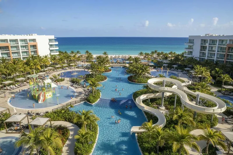 Large resort pool complex with palm trees, buildings, water slides, and the ocean in the background
