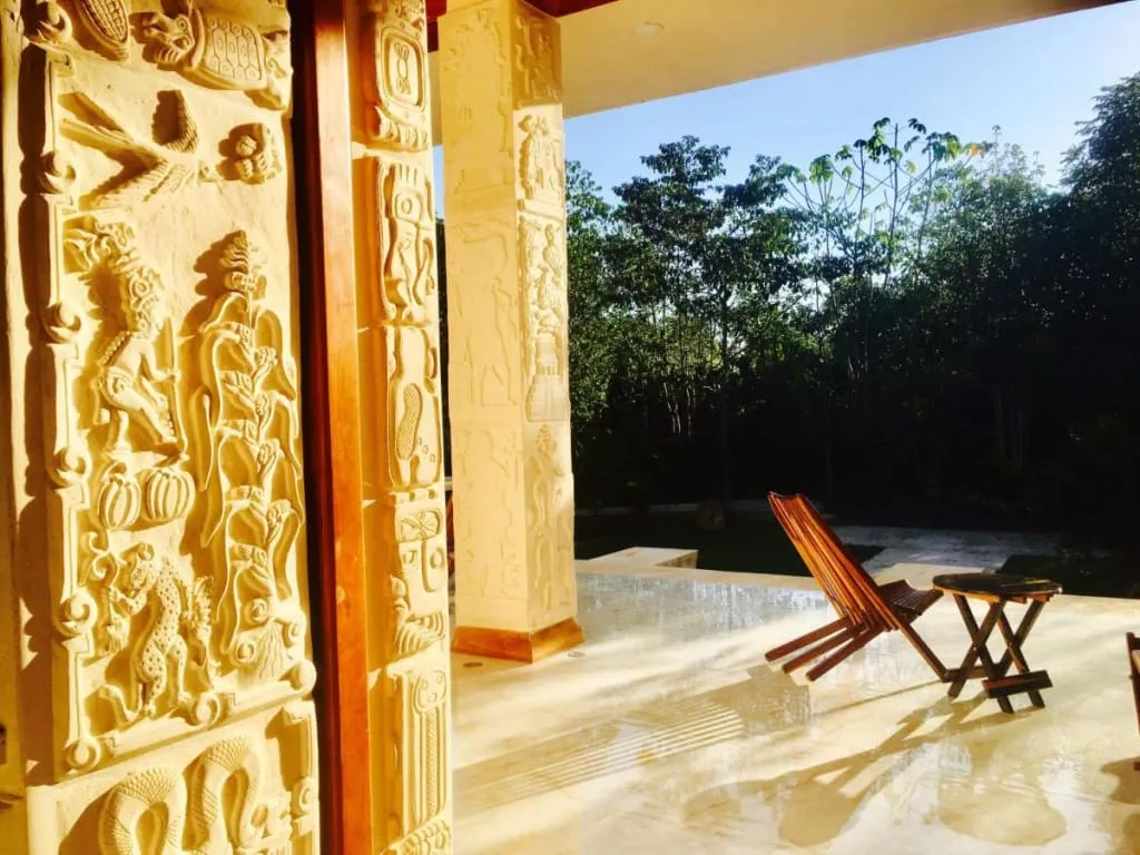 Sunlit patio with carved cream columns, two wooden chairs, and a small table beside trees