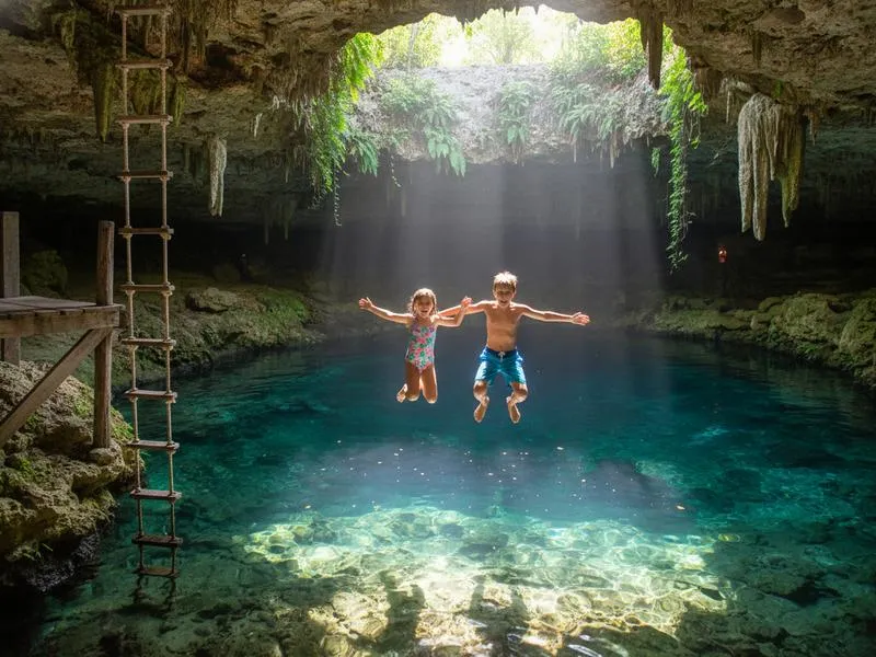 Children jumping into a natural cenote in Yucatan near Cancun, crystal clear turquoise water below, rocky limestone edges, bright sunlight, happy children in swimwear