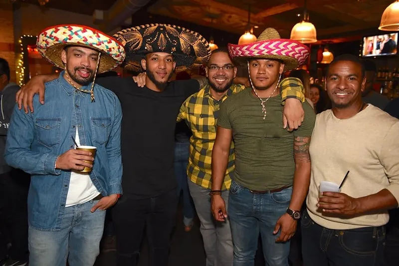 Five men posing together indoors, three wearing colorful sombreros and two holding drinks.
