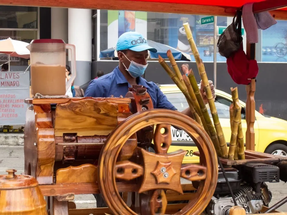 Trapiche hand-cranked sugarcane press at a Mexican market — vendor feeding stalks through rollers to extract guarapo juice