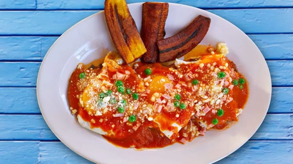 Huevos Motuleños served on tortillas with black beans, ham, peas, and plantain