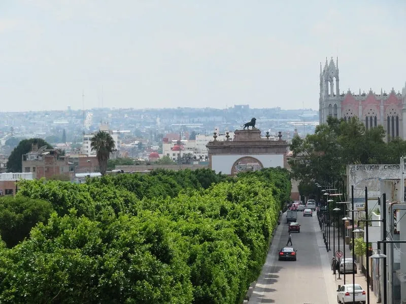 Shaded central plaza in Leon with benches, trees, and warm clouds before an afternoon shower