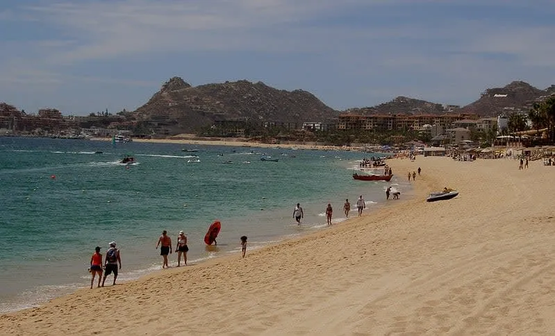 People walking along a sandy beach with mountains and waterfront buildings behind