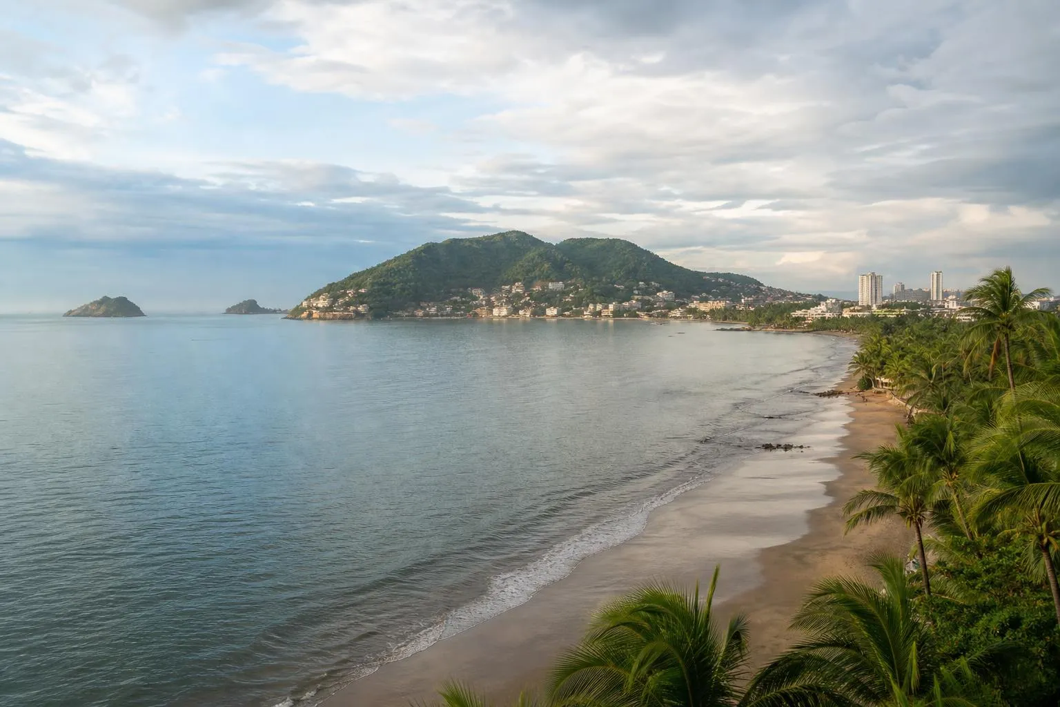 Manzanillo bay with palm-lined beach, green hills, and humid August clouds on Mexico's Pacific coast