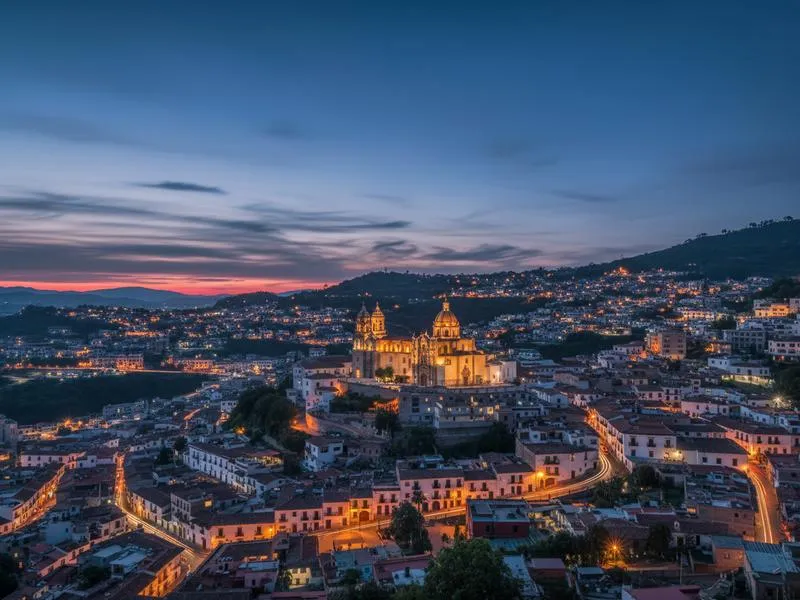Twilight blue hour photograph of Taxco Guerrero Mexico with white colonial buildings cascading down hillside and illuminated Santa Prisca church dome glowing warmly against moody sky