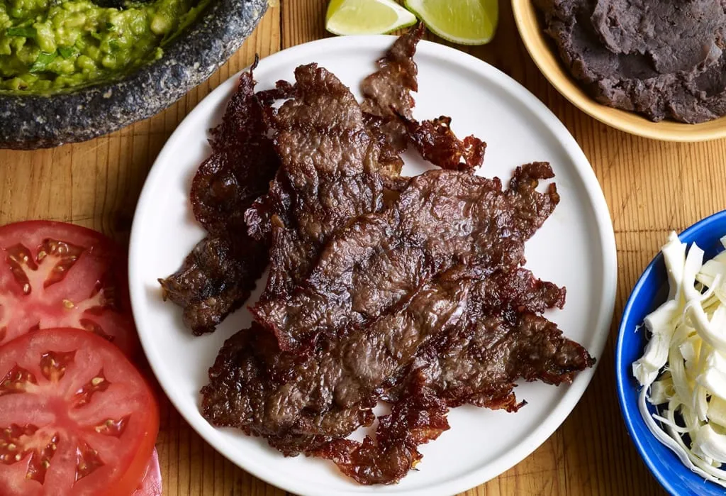 Oaxacan tasajo hanging to dry vertically near wood fires — the rope-rolled form and smoke give it a completely different character from Morelos cecina