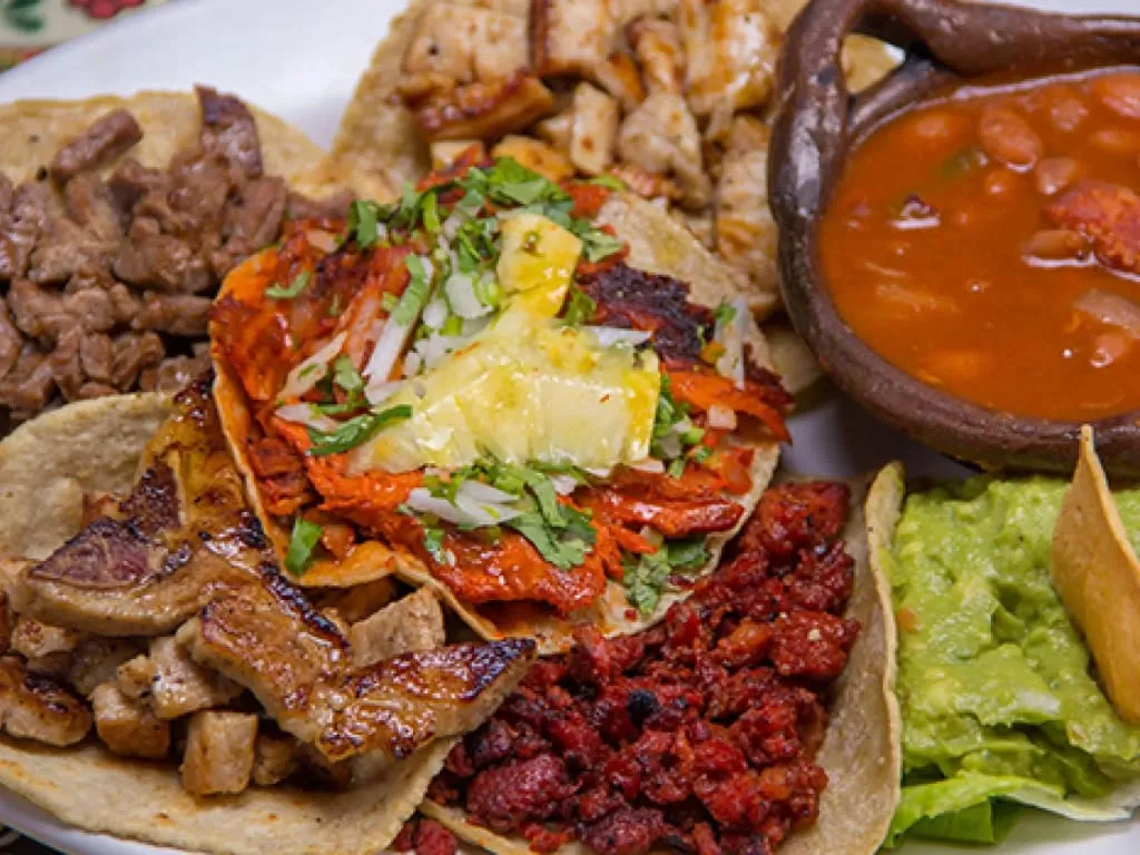 A plate holds several tacos topped with meat, pineapple, onion, and cilantro beside beans and guacamole