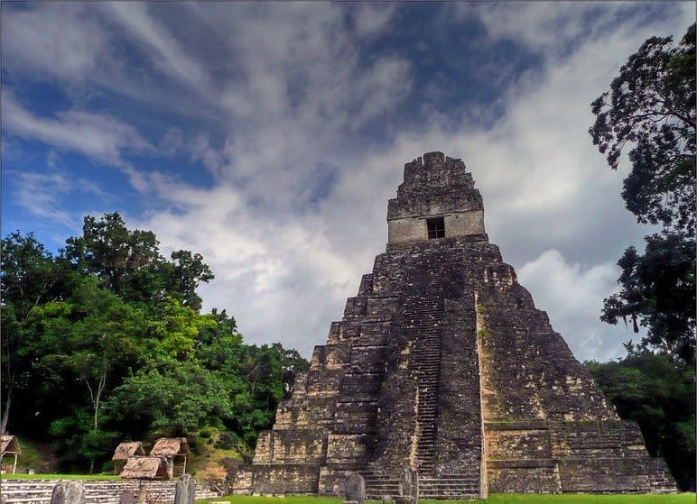 Tall steep stone pyramid in a clearing surrounded by trees beneath cloudy skies