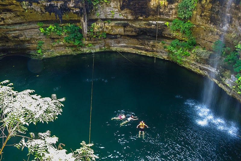 Swimmers in Cenote Saamal beneath a waterfall and hanging greenery