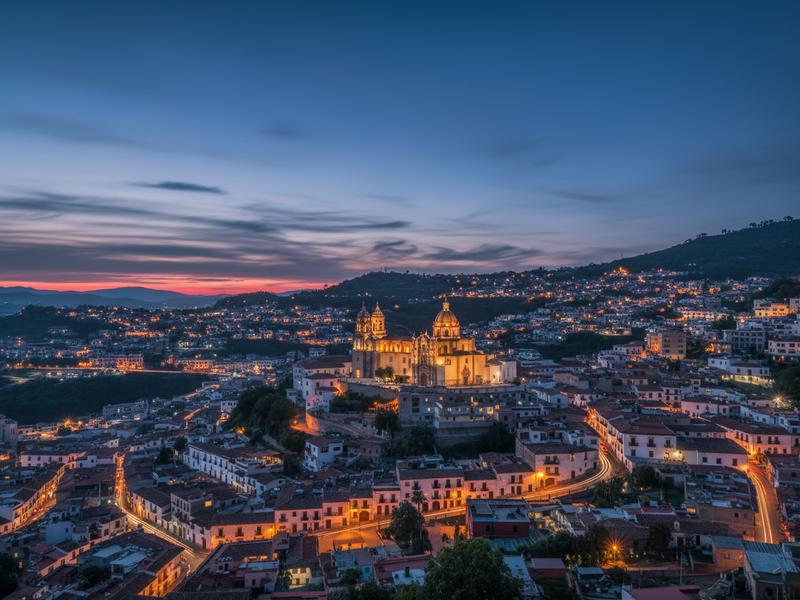 Twilight blue hour photograph of Taxco Guerrero Mexico with white colonial buildings cascading down hillside and illuminated Santa Prisca church dome glowing warmly against moody sky