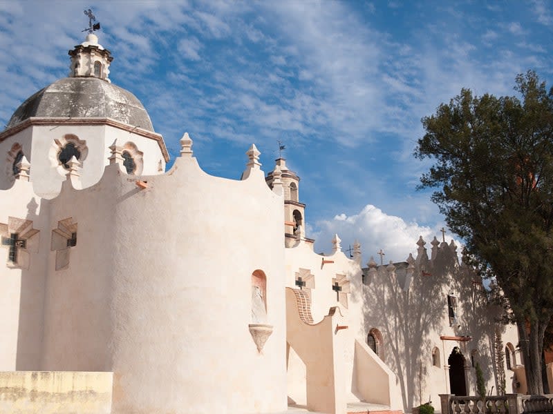 White church complex with a domed roof, sculpted walls, crosses, and trees under a blue sky