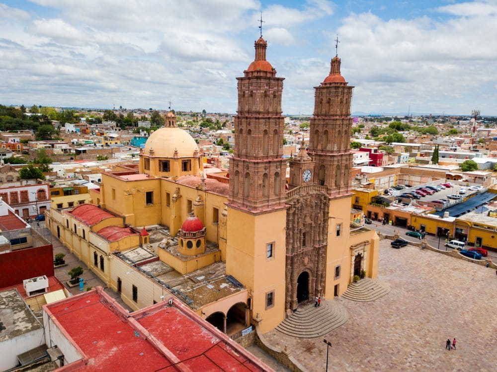 Aerial view of a large yellow cathedral with twin towers, domes, and a wide stone plaza