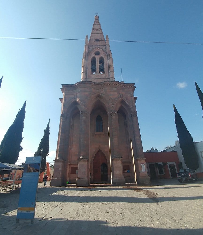 Tall narrow stone church facade with a central pointed entrance, bell tower, and cypress trees on either side