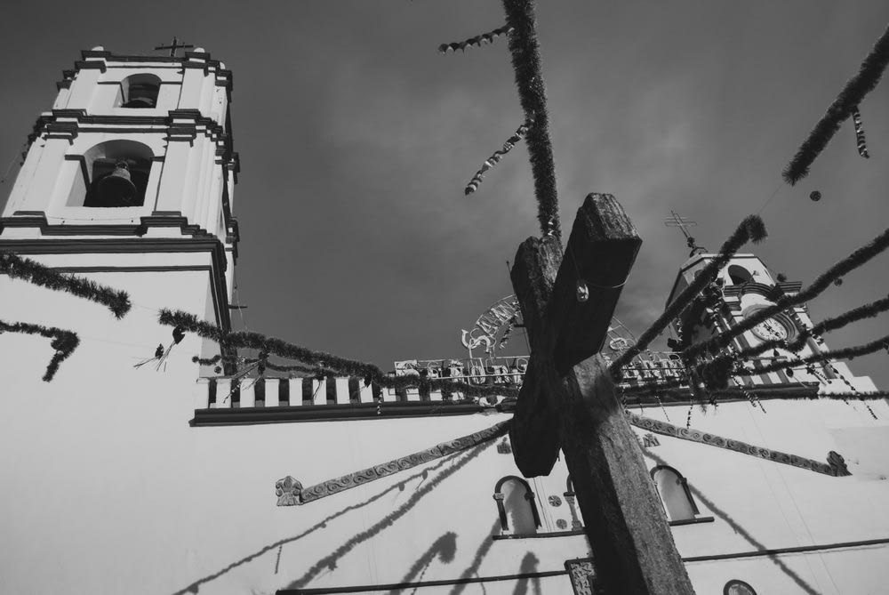 Black-and-white low-angle view of a church facade with two bell towers and a large wooden cross in the foreground