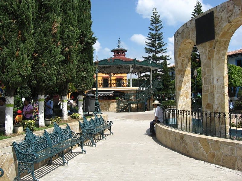Small town square with ornate metal benches, a gazebo, trees, and a stone arch structure