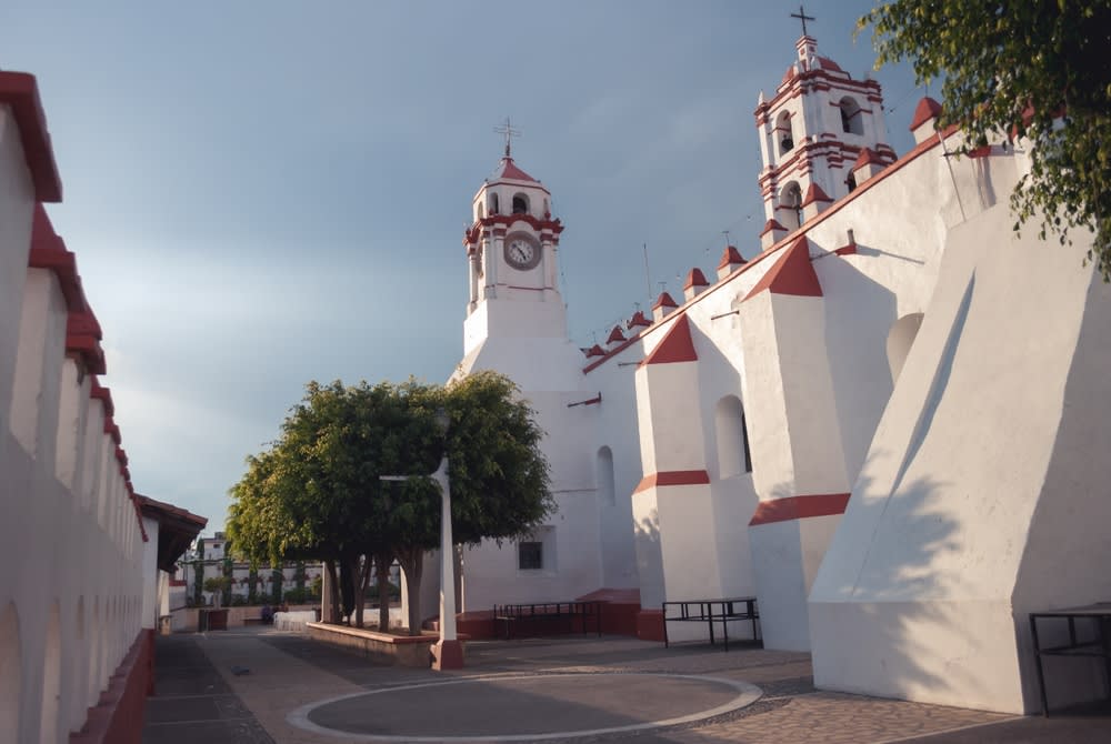 White church with red trim, clock tower, bell tower, and a narrow paved courtyard lined with trees