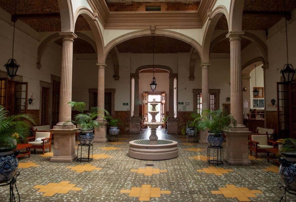 Interior courtyard with tall columns, arches, potted plants, chairs, and a small fountain on patterned tile flooring