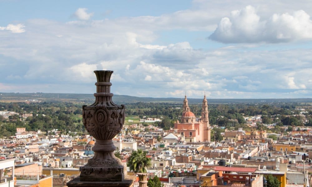 Cityscape viewed from above with an ornate stone urn in the foreground and a church with twin towers in the distance