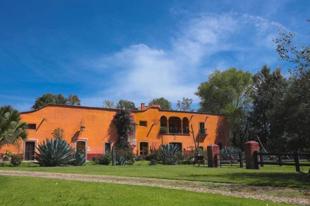 Long orange hacienda-style building beside a green lawn with agave plants, trees, and a blue sky