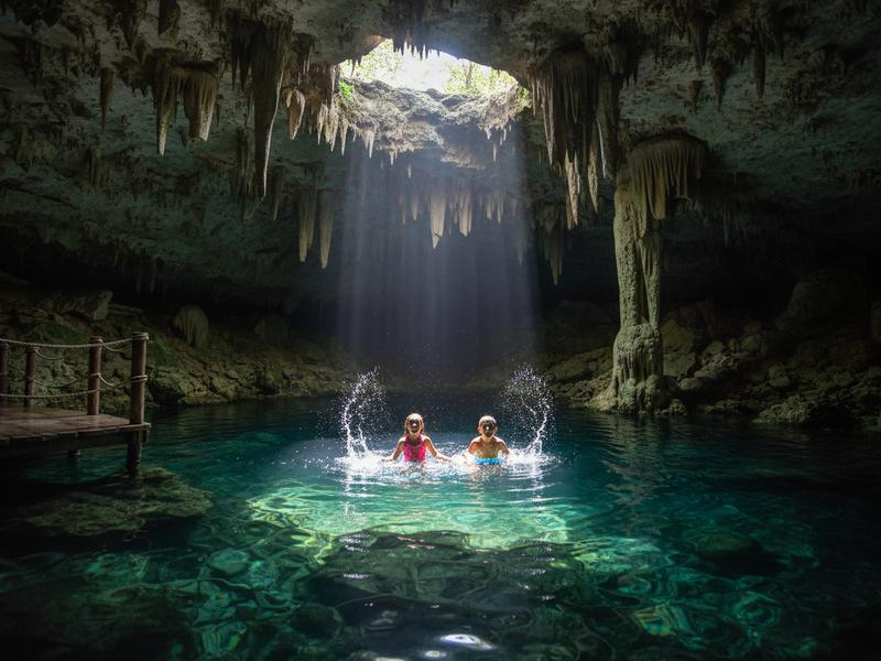 Children swimming in a clear turquoise cenote in Yucatan Mexico with stalactites above and a wooden platform for jumping