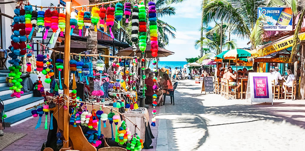 Beachfront lane lined with colorful hanging crafts, restaurants, palm trees, and ocean views