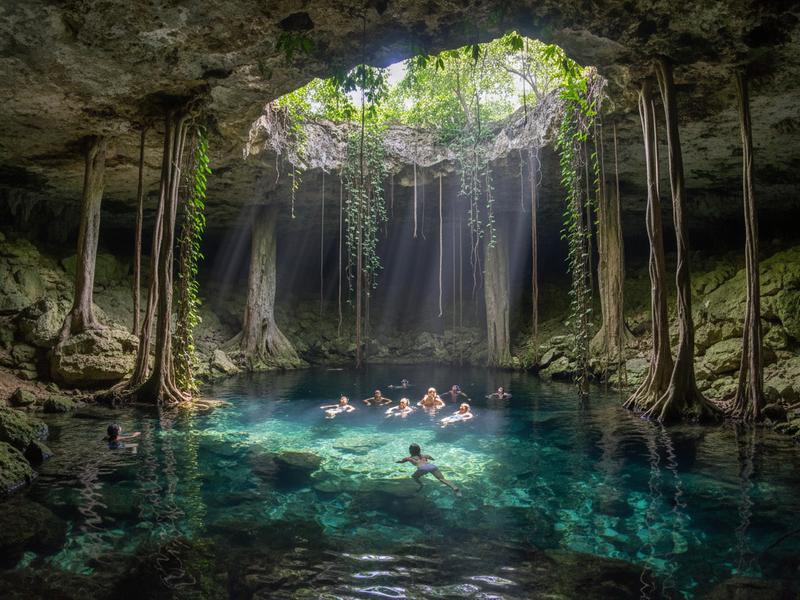 Travelers swimming in a turquoise cenote with limestone formations and hanging vines
